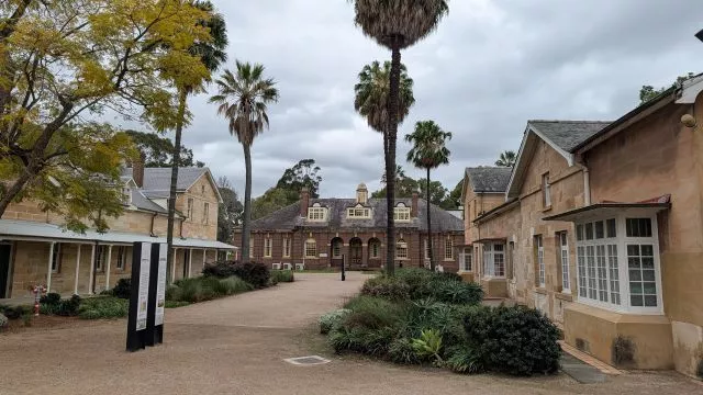Historic sandstone buildings, a brick facade building, and tall palm trees along the pathway of the Parramatta Female Factory precinct.
