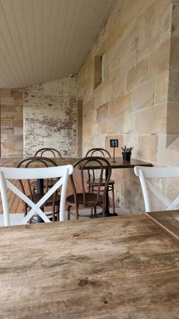 Wooden tables and chairs set up under the covered veranda against historic sandstone walls at Havenstone Cafe.