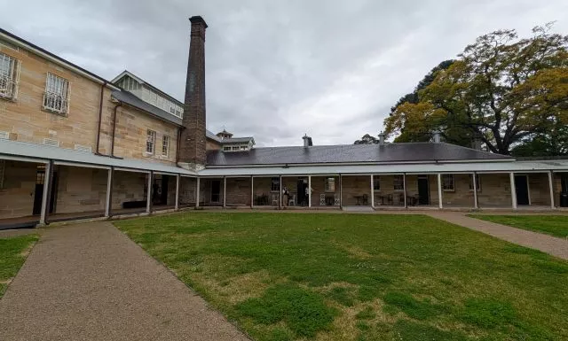 Historic sandstone buildings, a tall brick chimney, and a covered veranda surrounding the grass courtyard at Havenstone Cafe.