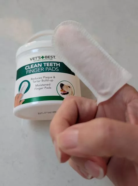 A hand wearing a white textured dental finger wipe on the index finger in front of a tub of Vet's Best Clean Teeth Finger Pads.