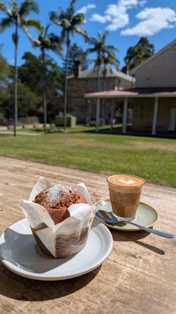 A freshly baked muffin and glass of latte on an outdoor timber table with the historic sandstone buildings of the Parramatta Female Factory precinct in the background