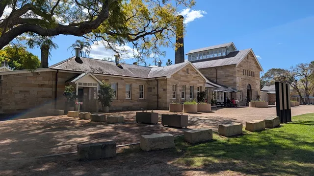 Wide view of the heritage sandstone buildings, tall chimney stack, outdoor courtyard seating, and leafy jacaranda branch at Havenstone Cafe.